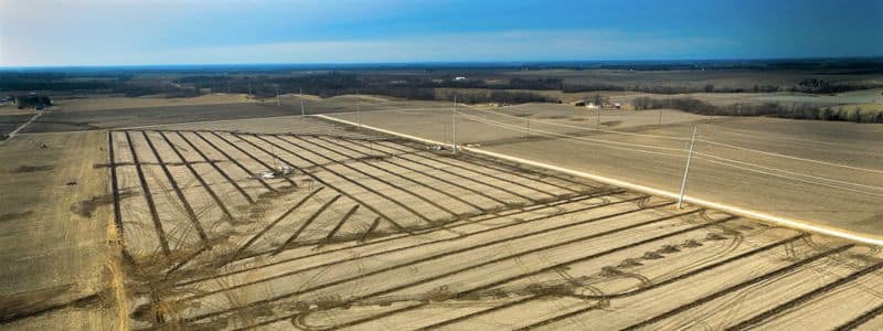 Aerial view of expansive flat farmland reveals parallel lines of tilled soil featuring a meticulous drainage tile design. Sparse trees dot the landscape beneath a clear blue sky in the background.