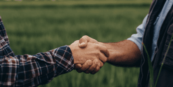 Two people shaking hands in a green field, their casual outdoor clothing reflecting a relaxed atmosphere. The scene captures the camaraderie of an outsourced accounting partnership built on trust and shared goals.