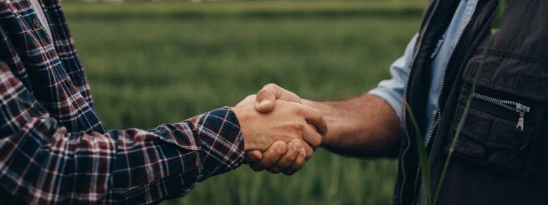Two people shaking hands in a green field, their casual outdoor clothing reflecting a relaxed atmosphere. The scene captures the camaraderie of an outsourced accounting partnership built on trust and shared goals.