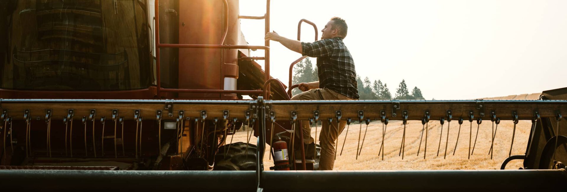 A person in a plaid shirt navigates the farm risks while climbing onto a large agricultural machine in a sunny field.