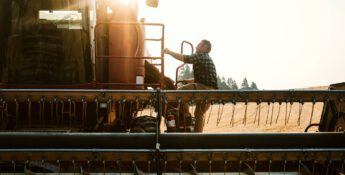 A person in a plaid shirt navigates the farm risks while climbing onto a large agricultural machine in a sunny field.