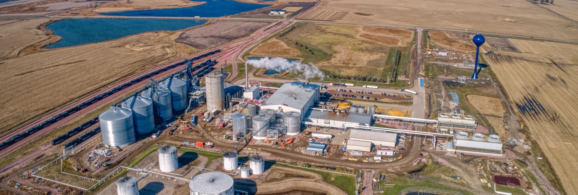 An aerial view reveals an industrial facility with multiple storage tanks eligible for the 45Z Tax Credit, surrounded by farmland. Nearby, a water tower and ponds accentuate the landscape, while fields stretch into the horizon under a clear sky.