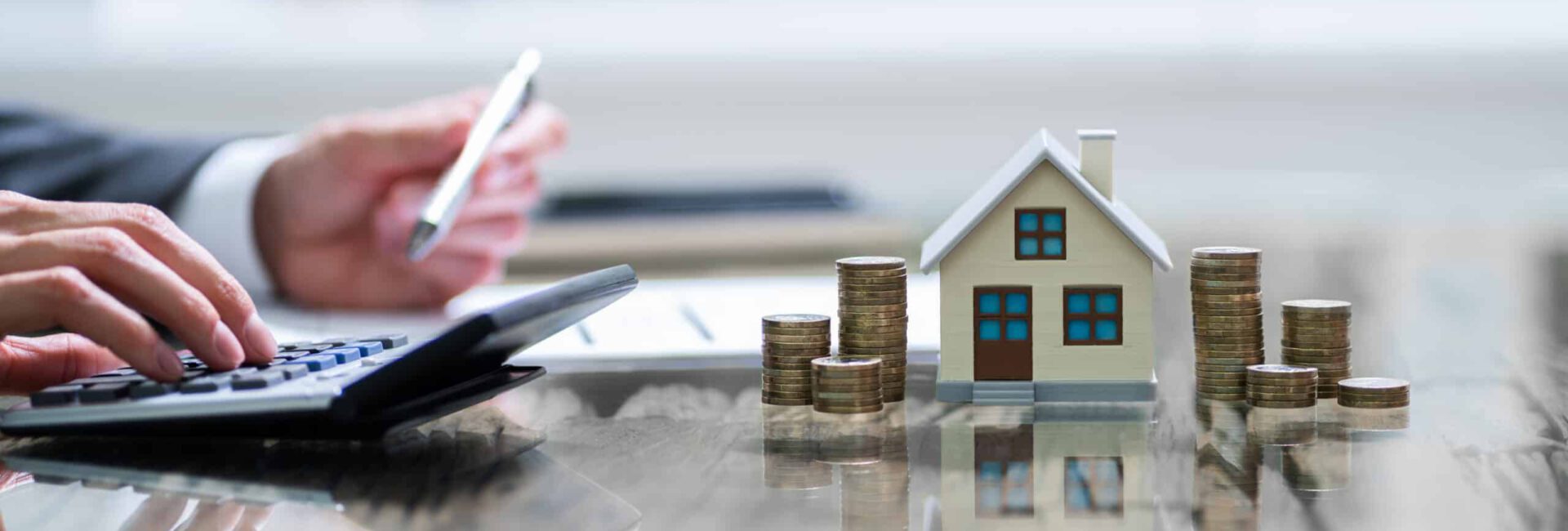 A person uses a calculator beside a small house model and stacks of coins on a reflective surface, illustrating financial planning or investment in real estate, possibly guided by HUD strategies.