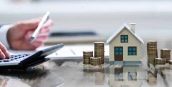A person uses a calculator beside a small house model and stacks of coins on a reflective surface, illustrating financial planning or investment in real estate, possibly guided by HUD strategies.