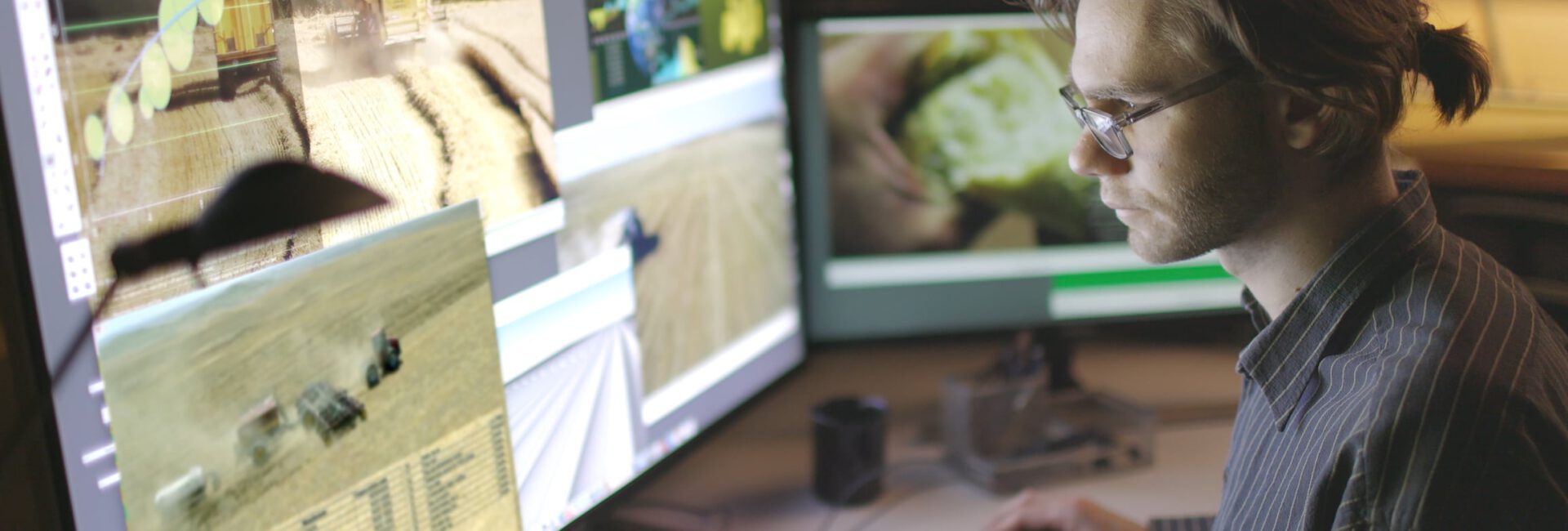 A person with glasses sits at a tech-savvy farm office desk, viewing multiple monitors displaying images of agricultural machinery and fields.