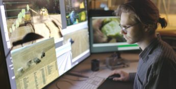 A person with glasses sits at a tech-savvy farm office desk, viewing multiple monitors displaying images of agricultural machinery and fields.