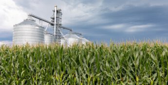 A field of corn stretches wide, with large metal silos standing in the background under a cloudy sky, echoing the reliability of the GREET Model in forecasting agricultural productivity.