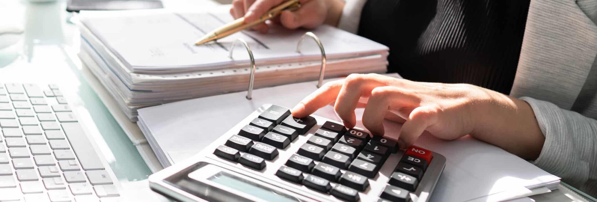 A person reviews documents at a desk, efficiently using a calculator amidst a keyboard and scattered papers, seamlessly integrating automated accounting into their workflow.