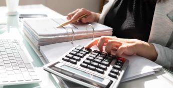 A person reviews documents at a desk, efficiently using a calculator amidst a keyboard and scattered papers, seamlessly integrating automated accounting into their workflow.