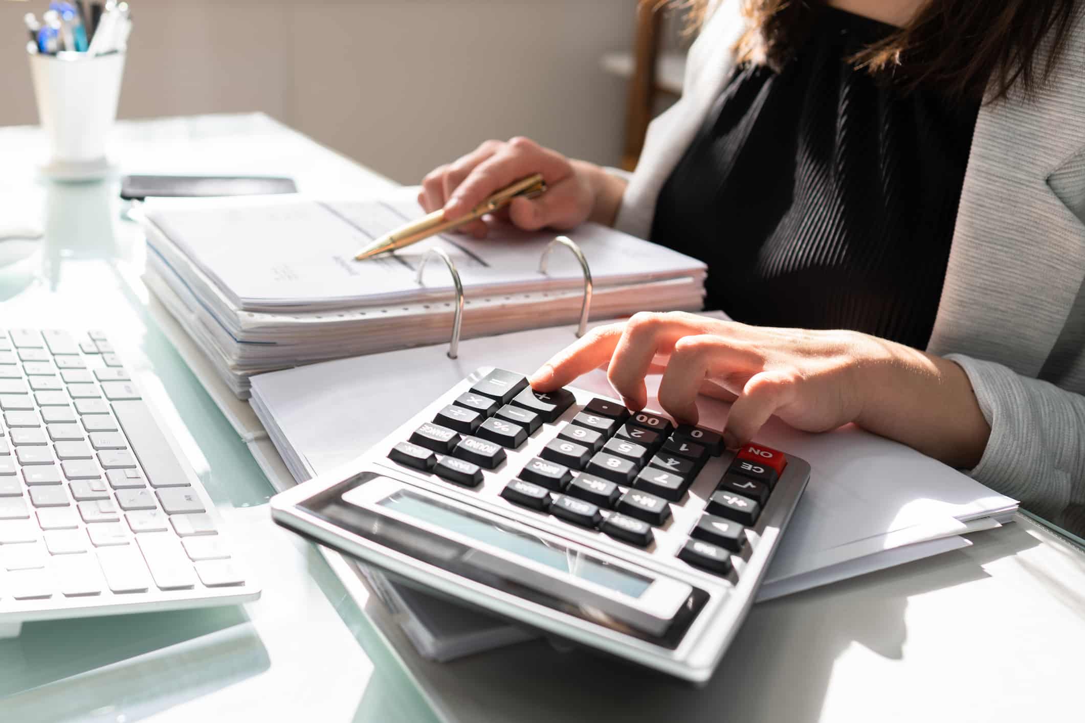 A person reviews documents at a desk, efficiently using a calculator amidst a keyboard and scattered papers, seamlessly integrating automated accounting into their workflow.