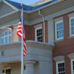 A brick and stone bank building with large windows proudly displays a U.S. flag out front, reflecting the steadfast nature of financial institutions amidst changing ag impacts.