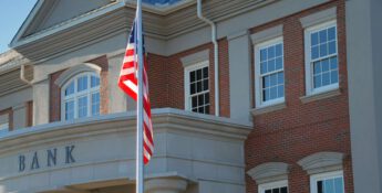A brick and stone bank building with large windows proudly displays a U.S. flag out front, reflecting the steadfast nature of financial institutions amidst changing ag impacts.