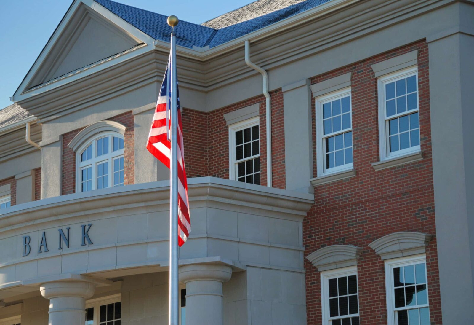 A brick and stone bank building with large windows proudly displays a U.S. flag out front, reflecting the steadfast nature of financial institutions amidst changing ag impacts.