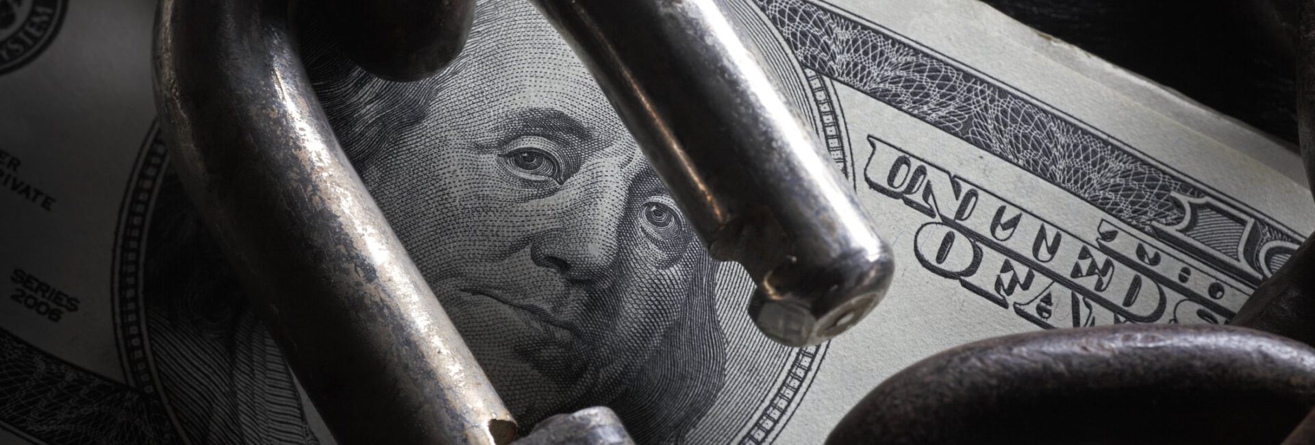 A hundred-dollar bill is partially visible beneath a chain and padlock, the lock open, symbolizing how to increase business wealth by overcoming financial restriction or enhancing security measures.