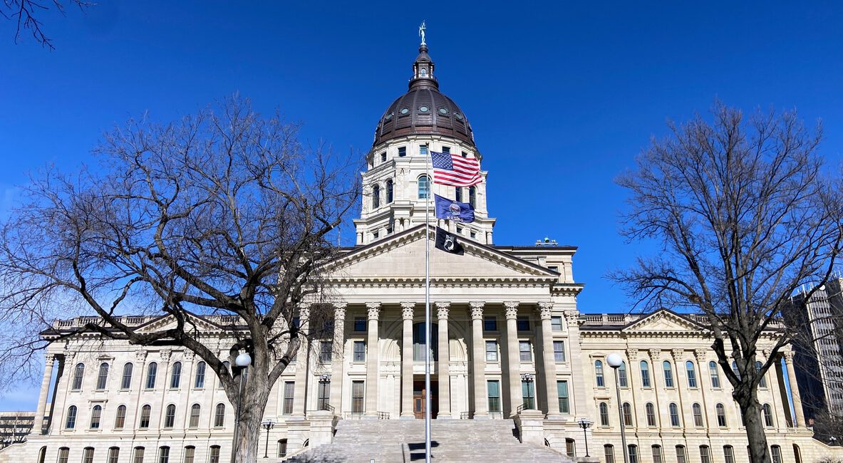 Front view of a large, historic building with a prominent dome and columns, surrounded by leafless trees. The American flag and another representing Kansas state income tax reforms fly in front. Clear blue sky provides the perfect backdrop for this iconic scene.