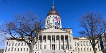 Front view of a large, historic building with a prominent dome and columns, surrounded by leafless trees. The American flag and another representing Kansas state income tax reforms fly in front. Clear blue sky provides the perfect backdrop for this iconic scene.