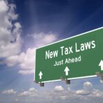 Green highway sign reads "Newly Proposed 2023 Tax Deductions Just Ahead" with arrows pointing forward, set against a partly cloudy sky.