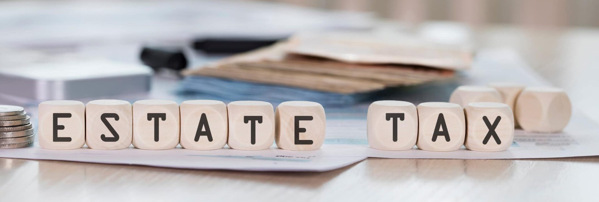 Lettered blocks spell "ESTATE TAX" on a table, surrounded by financial documents, coins, and hints of estate tax exemptions in the backdrop.