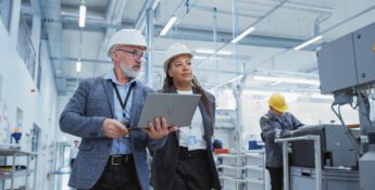 Two people in hard hats, one with a laptop, walking through a manufacturing facility. In the background, a worker operates machinery.