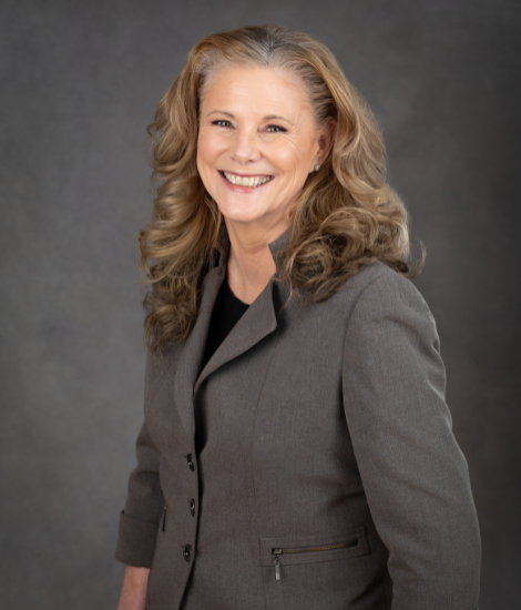 A woman with long, wavy hair wearing a gray business suit smiles against a gray background.