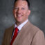 A man in a tan suit jacket, white patterned shirt, and red tie smiles at the camera against a gray background.