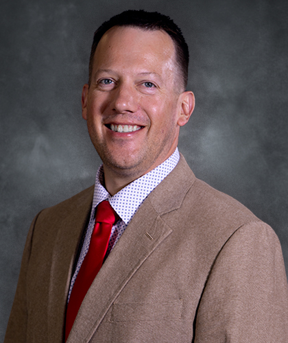 A man in a tan suit jacket, white patterned shirt, and red tie smiles at the camera against a gray background.