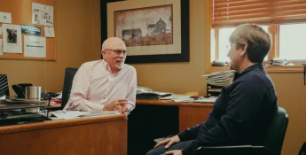 Two people are sitting and talking in an office. One is seated at a desk, and the other in a chair facing them. The office has papers, a picture frame, and a window with blinds.