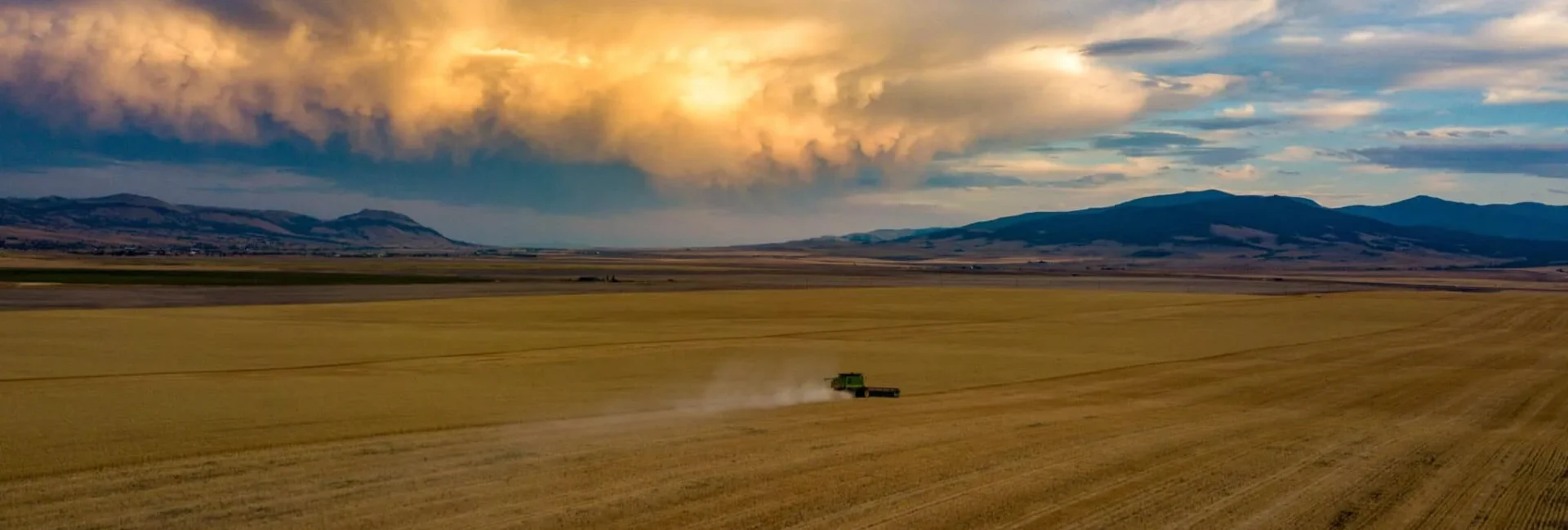 A vast field under a dramatic sky with scattered clouds and sunset hues. A combine harvester creates a dust trail as it moves across the landscape, with hills visible in the background.