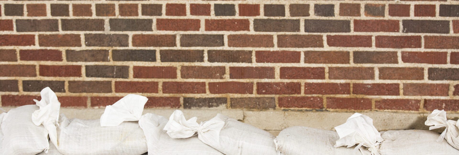 A row of white sandbags is placed in front of a red and brown brick wall on a sidewalk.