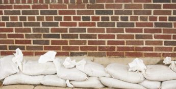 A row of white sandbags is placed in front of a red and brown brick wall on a sidewalk.