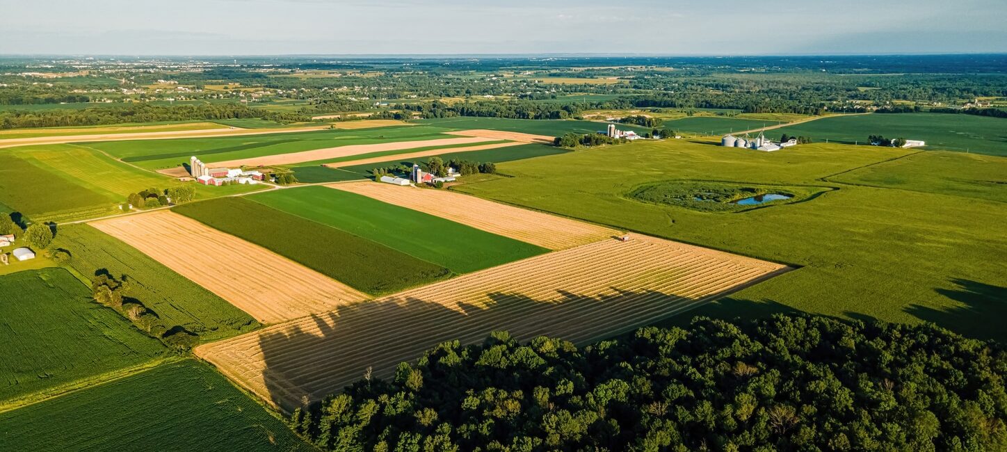 Aerial view of a rural landscape with agricultural fields, a few farm buildings, and patches of forest under a clear sky.