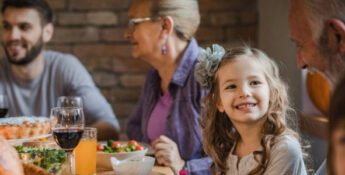 A family gathers around a table filled with food and drinks. A young girl smiles while sitting with adults.