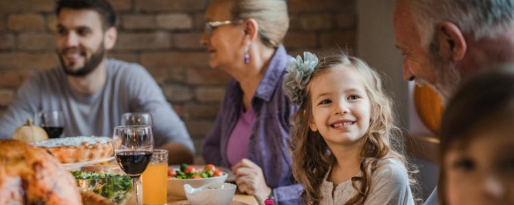 A family gathers around a table filled with food and drinks. A young girl smiles while sitting with adults.