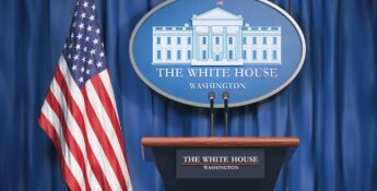 Press podium at the White House with microphones, a sign displaying "The White House, Washington," and a U.S. flag in the background against a blue curtain.