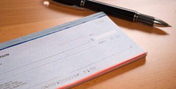 A checkbook with blank checks rests on a wooden surface, its potential for check fraud subtly offset by the presence of a black and silver pen.