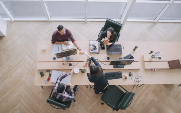 Four people sit at two connected desks, engaged in the office's workshare plan. Their computers hum softly as two of them share pastries, coffee cups scattered around. Large windows let in ample light, creating a collaborative and inviting atmosphere.
