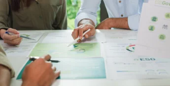 Three people reviewing ESG-related documents and graphs at a table, with one person pointing to a green chart with a pen.