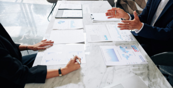 Two people sit at a table with charts and graphs on paper. One person gestures while speaking, and the other takes notes. A laptop and clipboard are also on the table.