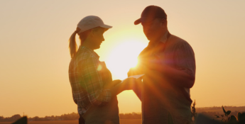 Two people in caps stand facing each other outdoors at sunset, with the sun glowing between them.