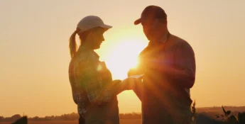 Two people in caps stand facing each other outdoors at sunset, with the sun glowing between them.