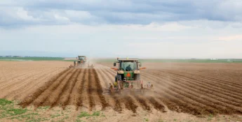 Two tractors plowing a vast, flat field under a cloudy sky, with soil turning up in neat, parallel rows.