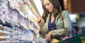 Woman in a green jacket and polka dot dress shops in a grocery store, examining a product from a refrigerated shelf while holding a shopping basket.