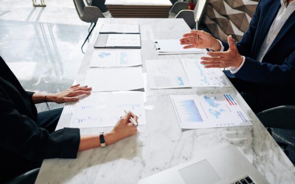Two people in a meeting analyzing charts and graphs on a marble table, with a laptop and a tablet nearby.