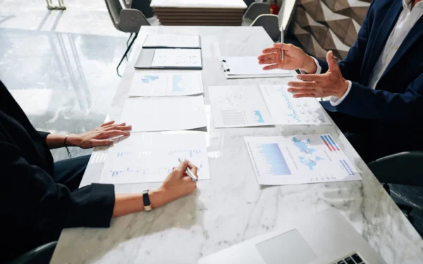 Two people in a meeting analyzing charts and graphs on a marble table, with a laptop and a tablet nearby.