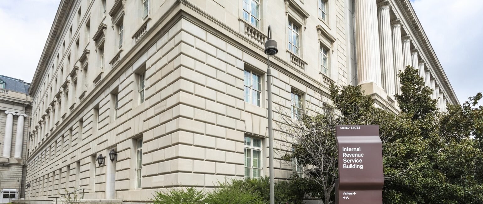 A large, beige stone building with multiple windows, identified by a nearby brown sign as the Internal Revenue Service Building.