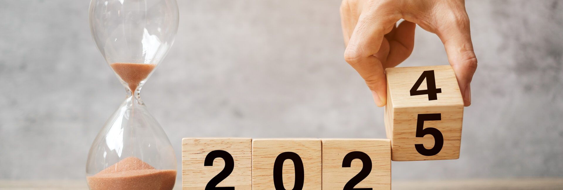 A hand flips a wooden block from 2024 to 2025, symbolizing the transition to a new year. An hourglass filled with sand sits beside it, reminding us of the importance of time management as part of essential HR year-end tips.