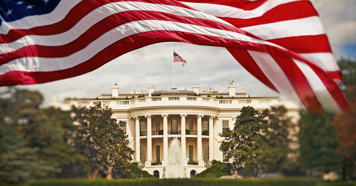 American flag waves in the foreground with the White House in the background, surrounded by greenery and a fountain.