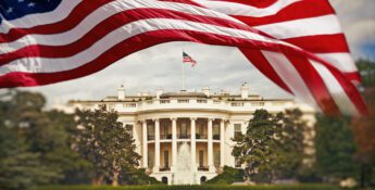 American flag waves in the foreground with the White House in the background, surrounded by greenery and a fountain.