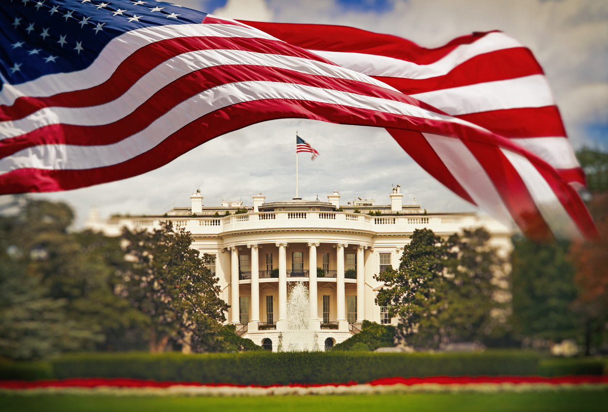 American flag waves in the foreground with the White House in the background, surrounded by greenery and a fountain.