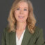 Becky Ringenberg, with shoulder-length blonde hair, wears a green blazer over a white blouse as she poses confidently in front of a plain dark background.